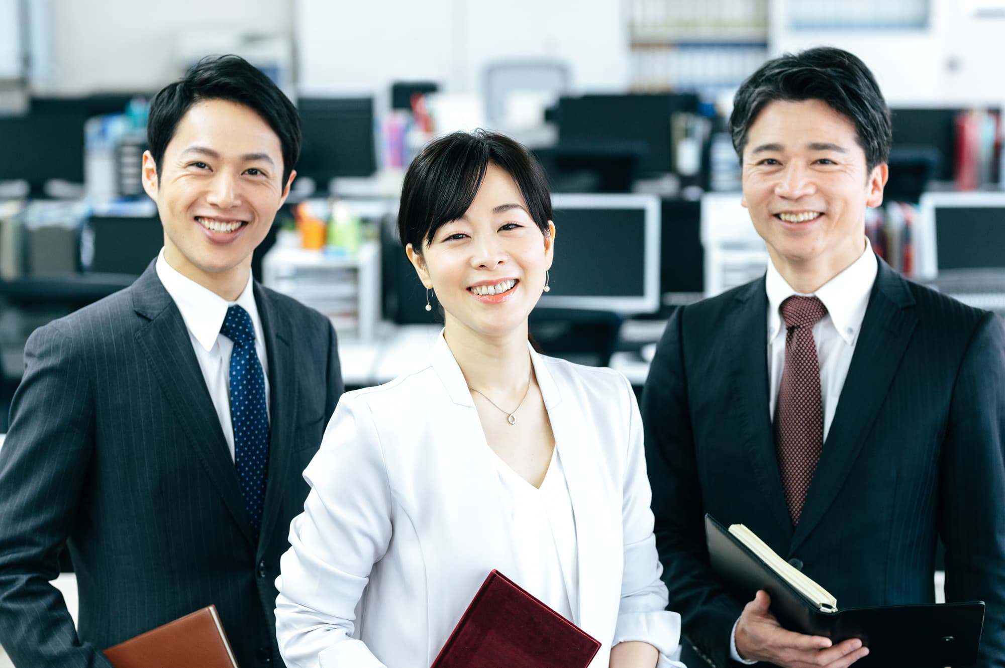パソコンが並ぶ社内で、手帳を携えてこちらに優しく微笑むスーツ姿の男女社員