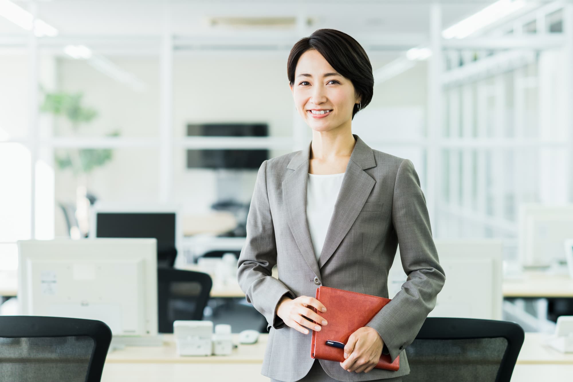 明るい社内で手帳を携え優しくこちらに微笑みかけるスーツ姿の女性